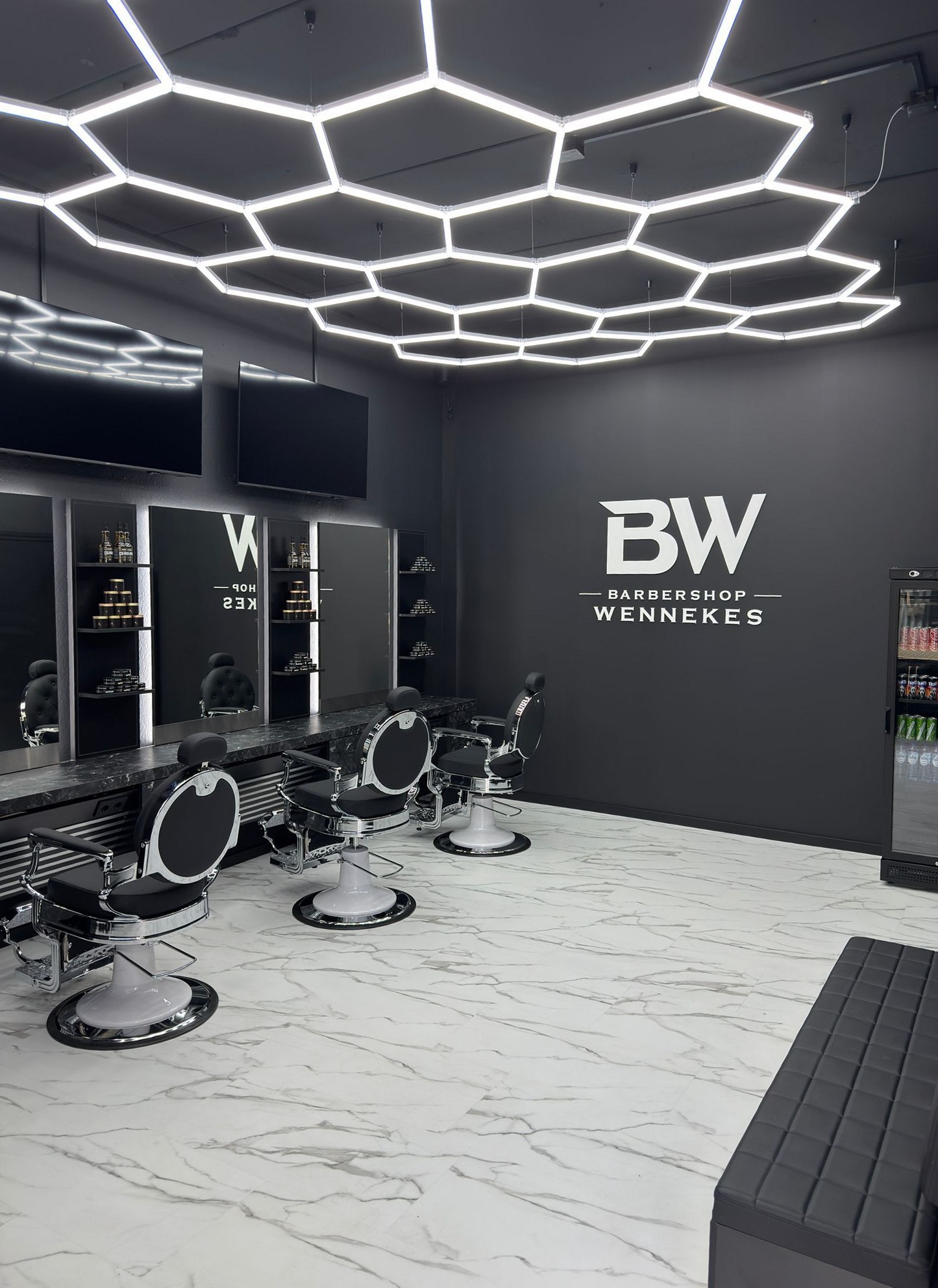 Binnen bij Barbershop Wennekes: drie barberstoelen voor de spiegels, de zwarte wand met het BW-logo, een witte marmeren vloer en het plafond van witte LED-zeshoeken