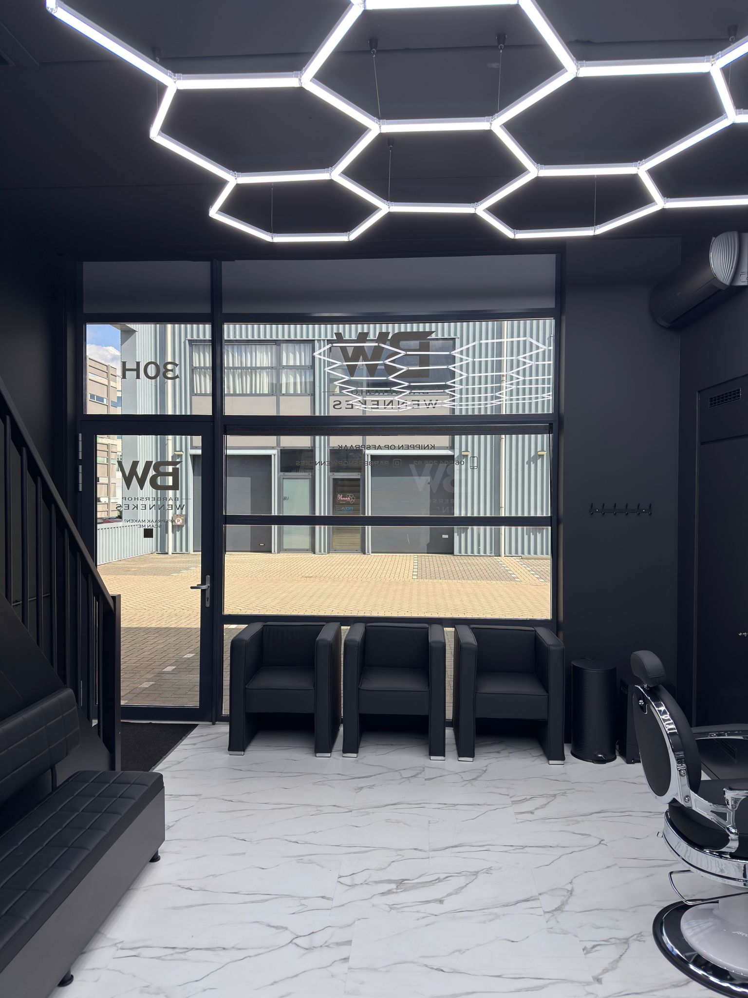 De wachtplek bij Barbershop Wennekes: zwarte stoelen voor de glazen pui, een barberstoel en het zeshoekige LED-plafond